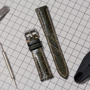 May include: A two-piece olive green watch strap featuring a textured, reptilian-like pattern and light tan contrast stitching. The strap includes a polished silver-tone metal buckle and two matching leather keepers, presented on a grid background.
