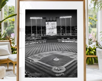 Può includere: Una fotografia in bianco e nero di uno stadio di baseball di notte. Lo stadio è illuminato da luci e il tabellone segnapunti mostra il nome e il numero di un giocatore. Il campo è vuoto e gli spalti sono per lo più vuoti. La parola "Royals" è visibile sul campo.