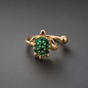 May include: A gold-toned open ring featuring a turtle-shaped centerpiece. The turtle shell is encrusted with multiple small, round, vibrant green stones. The ring band is smooth and polished, ending in a small gold sphere on one side.