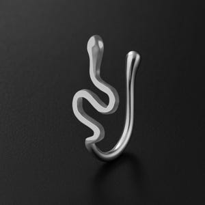 以下が含まれることがあります： 銀色のヘビの形をしたノーズカフ。ジュエリーは、ヘビのような頭と体を持つ湾曲したデザインです。カフは光沢のある金属でできており、暗い背景に配置されています。