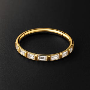 May include: A gold-colored ring with a series of rectangular, clear stones set around the band. The ring is on a black background, and the stones are evenly spaced around the ring's circumference.