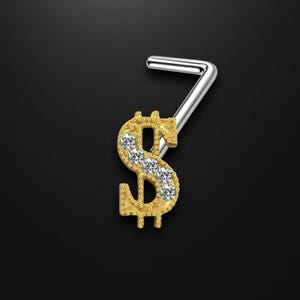 May include: A gold-colored nose ring in the shape of a dollar sign, embellished with clear gemstones. The nose ring has a silver-colored post. The design is set against a black background.