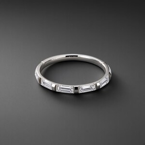 Könnte beinhalten: Ein silberfarbener Metallring mit einer Reihe klarer, rechteckiger Baguette-Schliff-Steine, die horizontal entlang des äußeren Umfangs eingesetzt sind. Der Ring hat eine glatte, polierte Oberfläche und einen sichtbaren Klappverschluss.