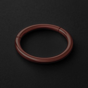 Peut inclure: Un anneau articulé circulaire de couleur cuivrée, probablement un bijou, est posé sur une surface sombre. L'anneau a une finition lisse et polie et un léger éclat métallique. La charnière est visible, indiquant qu'il peut s'ouvrir et se fermer. Le design est simple et moderne.