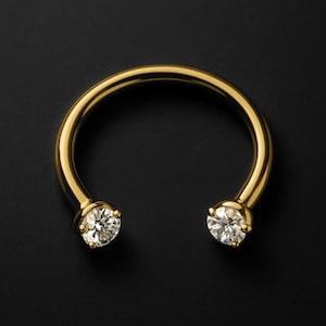 Peut inclure: Un anneau de piercing en forme de fer à cheval de couleur or, avec une pierre précieuse ronde et transparente à chaque extrémité. Le bijou est posé sur un fond noir, mettant en valeur sa surface polie et l'éclat des pierres.