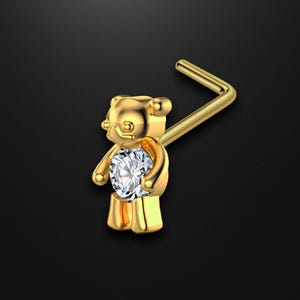 May include: A gold-colored nose stud featuring a teddy bear design. The bear holds a large, round, clear gemstone. The stud has a bent post for insertion. The background is a solid black.