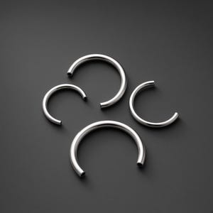 May include: Four polished silver-toned metal rings, each featuring a C-shaped open design with smooth, rounded ends. The set includes two larger rings and two smaller rings, all displaying a reflective, metallic finish with a consistent tubular shape.