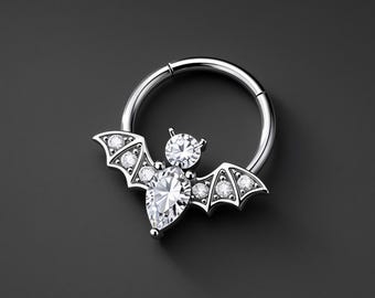 Puede incluir: Un anillo circular plateado con un dije en forma de murciélago. El murciélago está adornado con una gran piedra preciosa transparente en forma de pera para el cuerpo, una piedra redonda para la cabeza y tres pequeñas piedras en cada ala.