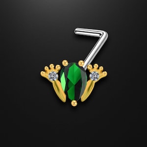 May include: A nose ring with a gold-coloured frog design, featuring a large, emerald-green gemstone. The frog's feet are decorated with small, clear gemstones. The nose ring has a silver-coloured, L-shaped post.