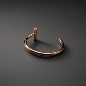 Può includere: Un bracciale rigido in oro rosa lucido caratterizzato da una fascia circolare liscia aperta a un'estremità. Un singolo montante cilindrico verticale è fissato su un lato dell'apertura, creando una distinta silhouette a forma di T con finitura riflettente.