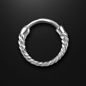 Könnte beinhalten: Ein silberner Nasenring mit einem gedrehten Seilmuster. Der kreisförmige Ring hat einen Klappverschluss und ist vor einem schwarzen Hintergrund platziert. Das gedrehte Design verleiht dem Schmuck eine einzigartige Textur.
