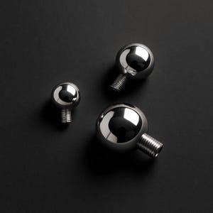 Op de afbeelding: Drie glanzende, verchroomde bollen met schroefdraadstelen liggen op een donkere achtergrond. De bollen, in verschillende maten, reflecteren licht. De schroefdraadstelen suggereren een mechanische of industriële toepassing.