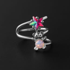 Peut inclure: Bague argentée avec un motif d'étoile et d'ours en peluche. L'étoile a une finition irisée multicolore, et l'ours en peluche tient une pierre opalescente ronde. La bague a un design en spirale.