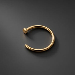 May include: A polished gold-tone circular hoop ring featuring a smooth, continuous band with a small, flat, cylindrical end cap. The jewellery piece has a reflective metallic finish and a simple, open-ended design that forms a nearly complete circle.