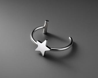 May include: A silver-toned metal ring featuring a smooth, five-pointed star charm attached to a thin, open-ended band. The ring has a polished, reflective surface and a small cylindrical post at one end of the opening, creating a minimalist design.