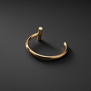 Op de afbeelding: Een gepolijste goudkleurige metalen manchetarmband met een gladde, ronde band die in een open ontwerp buigt. Het ene uiteinde van de armband eindigt in een korte, verticale cilindrische stijl, terwijl het andere uiteinde open en afgerond blijft.