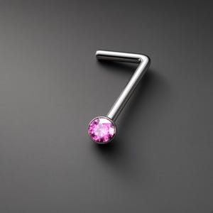 May include: An L-shaped nose stud featuring a polished silver-tone metal post and a single round, faceted pink gemstone set in a circular bezel. The jewelry piece has a smooth, reflective finish and a clear, vibrant pink stone at the end.