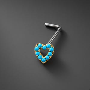 Puede incluir: Un piercing de nariz en forma de L con un engaste dorado en forma de corazón. El corazón está delineado con pequeñas piedras redondas de color turquesa brillante. El poste es de un material metálico plateado liso, doblado en ángulo recto.