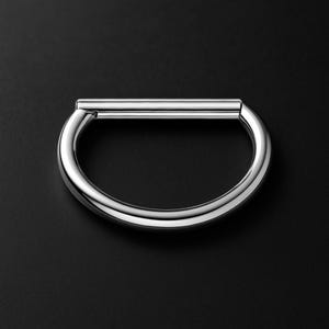Könnte beinhalten: Ein poliertes, silberfarbenes Metallarmband mit D-förmigem Design. Das Armband hat einen abgerundeten Boden und einen geraden oberen Balken, was eine elegante, moderne Ästhetik schafft. Das Metall reflektiert das Licht und hebt seine glatte Oberfläche vor dunklem Hintergrund hervor.