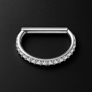 Puede incluir: Un aro de septum plateado, articulado, adornado con una hilera de piedras preciosas claras. La parte curva está engastada con piedras brillantes. La joya se presenta sobre un fondo oscuro, resaltando su superficie pulida y el brillo de las piedras.