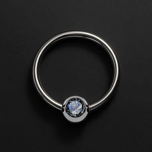Op de afbeelding: Een zilverkleurige captive bead ring met een ronde, heldere, blauwe edelsteen in het midden. De sieraden zijn cirkelvormig en hebben een gepolijste afwerking. De achtergrond is effen zwart.