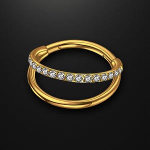 以下が含まれることがあります： ゴールドカラーのダブルフープピアス。上部のフープには、小さな透明な宝石が並んでいます。黒い背景に配置され、光沢のある反射面が強調されています。シンプルでありながらエレガントなデザインです。