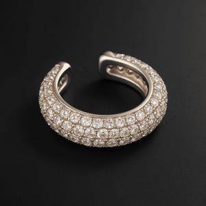 May include: A silver ear cuff encrusted with numerous small, sparkling diamonds. The cuff curves around the ear, with an open design at the top and bottom. The jewelry is set against a black background.