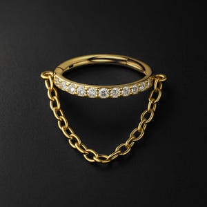 Könnte beinhalten: Ein goldfarbener Klappring mit einer Reihe kleiner, klarer Edelsteine. Eine zarte Goldkette hängt von dem Ring herab und erzeugt einen hängenden Effekt. Der Schmuck ist vor einem dunklen Hintergrund platziert.