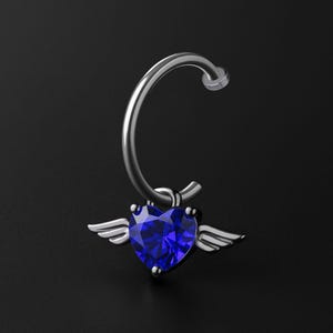 May include: A silver nose ring with a heart-shaped, faceted, deep blue gemstone and small silver wings. The ring has a curved design. The background is a solid black.