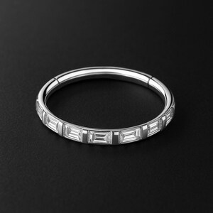May include: A silver ring with a continuous row of rectangular, clear stones. The ring is set against a black background, highlighting the sparkle of the stones. The ring is a piece of jewelry.