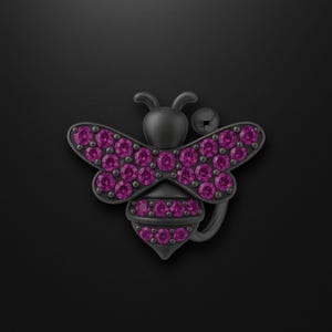 May include: A dark gray bee-shaped belly button ring with magenta-colored gemstones. The bee's wings and body are adorned with the gemstones, creating a sparkling effect. The jewelry is set against a black background.