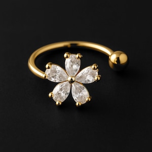 Puede incluir: Un anillo dorado con un diseño floral con cinco gemas claras en forma de lágrima. El anillo tiene una banda curva con una pequeña bola dorada en el extremo. El centro y los pétalos de la flor están acentuados con pequeños detalles dorados.