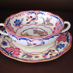 May include: A white porcelain soup bowl with two handles and a matching saucer. The set features a floral and bird design in pink, blue, and yellow, with a scalloped edge and gold trim. The bowl sits on the saucer.