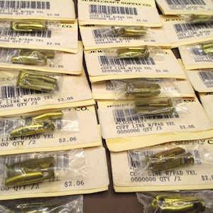 May include: A collection of gold-colored cuff links, each packaged in a clear plastic bag and attached to a cream-colored card. The cards have text and a barcode. The cuff links are a hardware supply.