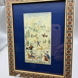 May include: A rectangular framed artwork featuring a miniature painting of a hunting scene with figures on horseback and animals in a landscape. The frame has an intricate geometric inlay pattern in red, black, and white, set against a dark blue mat.