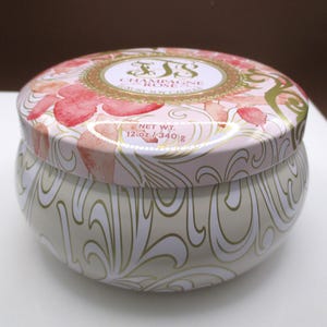 May include: A round, lidded candle tin with a floral design. The lid features pink strawberry illustrations and gold accents. The body of the tin is cream-colored with a gold, swirling pattern. Text on the lid reads "Champagne Rose" and "Luxury Soy Candle". Net wt. 12 oz / 340 g.