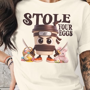 Op de afbeelding: Crèmekleurig T-shirt met de tekst "STOLE YOUR EGGS". De afbeelding toont een ninja-personage in de vorm van een koffiekopje met zwaarden, een mand met paaseieren en een roze konijn. Het personage draagt een hoofdband en oranje schoenen.