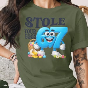 Op de afbeelding: Olijfgroen t-shirt met de tekst "STOLE YOUR EGGS". De afbeelding toont het cartoonnummer 67 met een lachend gezicht, witte handschoenen en schoenen, naast een mand met eieren en bloemen.