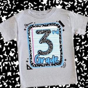 May include: A gray t-shirt featuring a notebook design with the words "3rd Grade" in a playful font. The design includes a blue and pink lined paper, scissors, a ruler, and a paperclip.