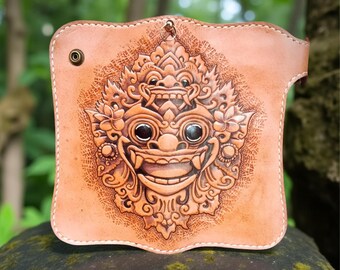 Könnte beinhalten: Hellbraune Ledergeldbörse mit geprägtem Design einer balinesischen Barong-Maske. Die Geldbörse hat einen Druckknopfverschluss und eine Schlaufe zum Aufhängen. Die detaillierte Schnitzerei zeigt florale und geometrische Muster.