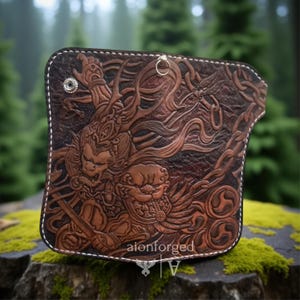 Puede incluir: Cartera de cuero marrón con intrincadas ilustraciones en relieve que representan una criatura mítica con una espada y detalles de cadenas. La cartera tiene un borde cosido blanco y un anillo de metal. El texto "aionforged" es visible.