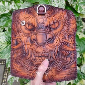 Pode incluir: Carteira de couro castanho com um design entalhado detalhado de uma máscara oni feroz. A carteira tem um anel em D na parte superior e um fecho de pressão. A marca "alonforged" é visível na parte inferior. Em um fundo de folhagem verde.