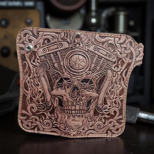 May include: A brown leather wallet with an embossed design featuring a skull, engine, and Celtic knot patterns. The wallet is rectangular with rounded corners and metal rivets.