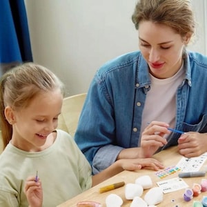 May include: A woman and a young girl are seated at a wooden table, engaged in a painting activity. The table holds white stones, small paint pots in various colors, paintbrushes, and a color palette. Both are focused on their artwork.