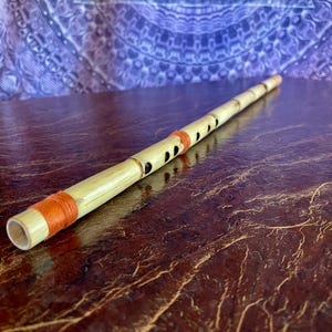 May include: A bamboo flute with a natural finish, orange bands and small holes. The instrument rests on a dark, textured surface, with a patterned purple backdrop. The flute is approximately 40 cm long.