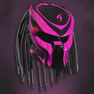 Puede incluir: Casco de moto negro y rosa con diseño Predator. El casco presenta un acabado negro brillante con detalles rosas y rastas. Se puede ver una luz roja en el lado izquierdo.