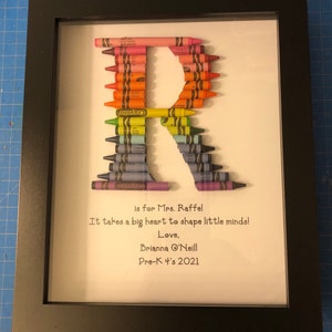 May include: A framed artwork featuring the letter "R" made from colorful crayons. The text "is for Mrs. Raffel It takes a big heart to shape little minds! Love, Brianna 'Neill Pre-K 4's 2021" is written below the letter.