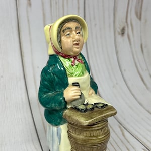 May include: A ceramic figurine of a woman in a green jacket and white apron, holding a bottle and oysters. The figurine is standing next to a small barrel. The base of the figurine has the words "Oysters! Twelve" written on it.