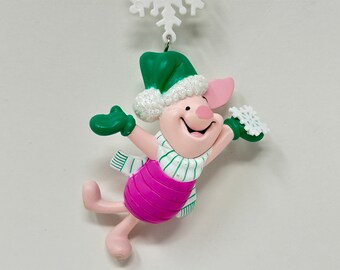 Puede incluir: Un cerdo rosa con un gorro y una bufanda verdes, sosteniendo un adorno de copo de nieve blanco. El adorno cuelga de una cuerda dorada con un copo de nieve blanco en la parte superior.