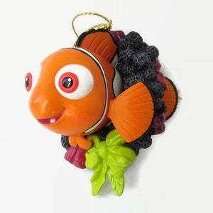 Peut inclure: Ornement de Noël en forme de poisson-clown orange et blanc avec une couronne noire, violette et verte. Le poisson sourit la bouche ouverte et a de grands yeux ronds.